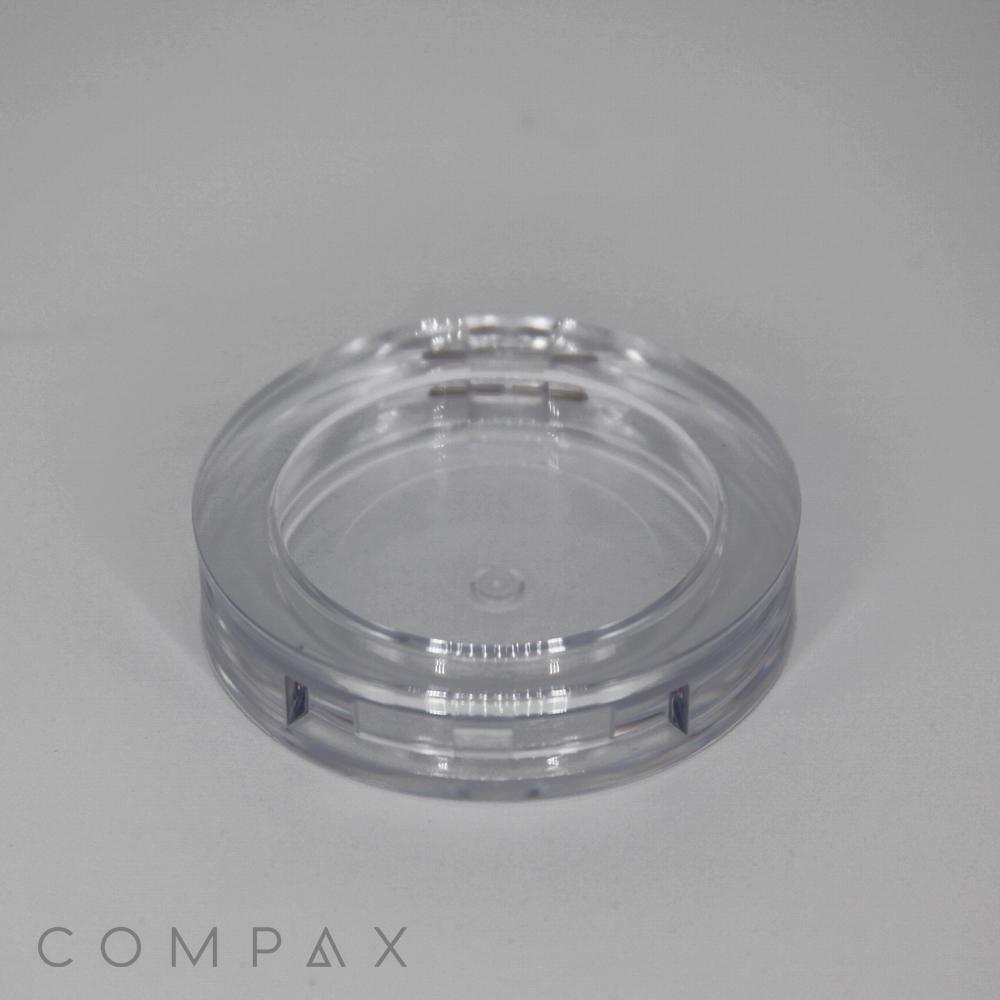 A transparent, round plastic container, named the Single Well Round Compact, with a clear lid sits on a gray surface. The container looks empty, and the word "COMPAX" is visible in the bottom left corner of the image.