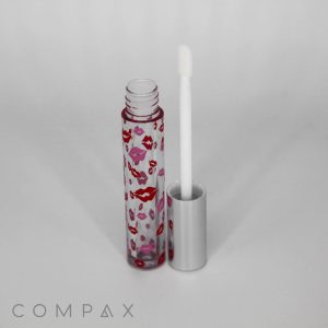 A 5mL Lip Gloss Bottle is shown with its cap removed and the applicator wand resting beside it. The tube has a clear design decorated with small red lips, while the cap boasts a sleek silver color. The brand name, COMAPX, is displayed in the bottom left corner.
