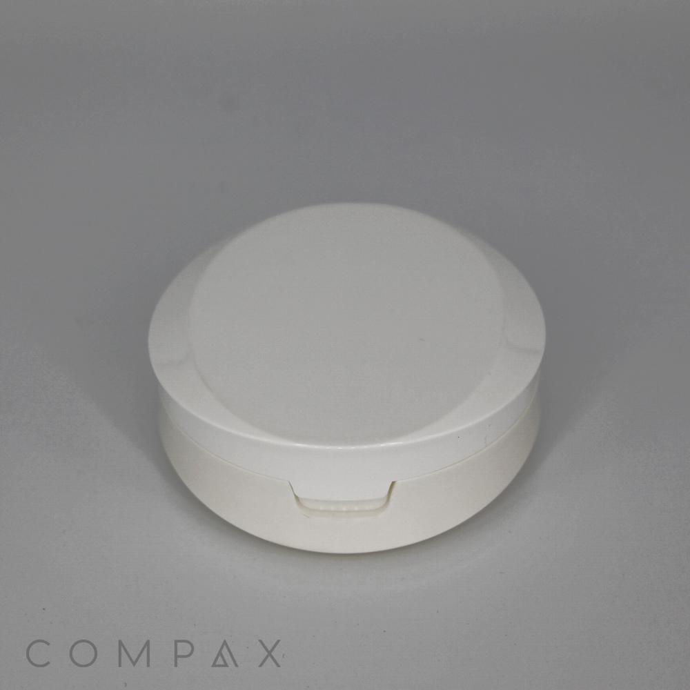 A round, white plastic Round Cushion Compact with Mirror featuring a snap-close lid is centered against a gray background. The word "COMPAX" is printed near the bottom left corner of the image in a minimalist font.