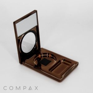 A Single Well Rectangular Compact in brown with an open lid, featuring a mirror on the inside. The base has circular and rectangular compartments for makeup products, and the word "COMPAX" is displayed in the bottom left corner of the image.