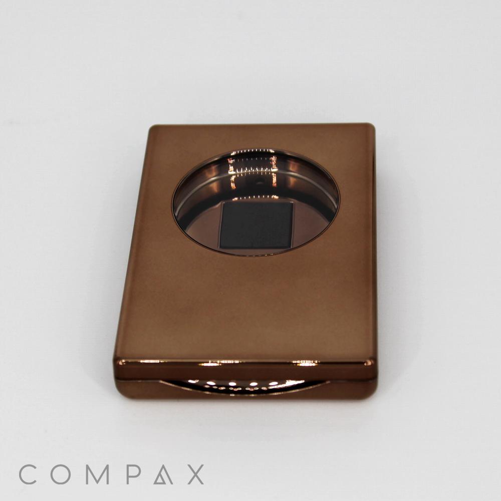 A sleek, metallic brown container named "Single Well Rectangular Compact" features a circular, transparent window at its center, revealing a small square object inside. The background is white, and the brand name "COMPAX" is displayed in light grey text at the lower left corner.