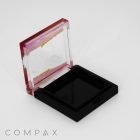 A square, open, empty Single Well Square Compact with a transparent pink lid and a black base. The lid is slightly angled, and the compact is positioned against a plain white background. The brand name "COMPAX" is visible at the bottom left corner.