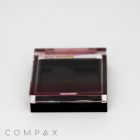 A closed Single Well Square Compact, featuring a black and transparent plastic design, reveals a partially visible pink substance inside. The brand name "COMPAX" is displayed at the bottom left corner of the image, set against a plain white background.
