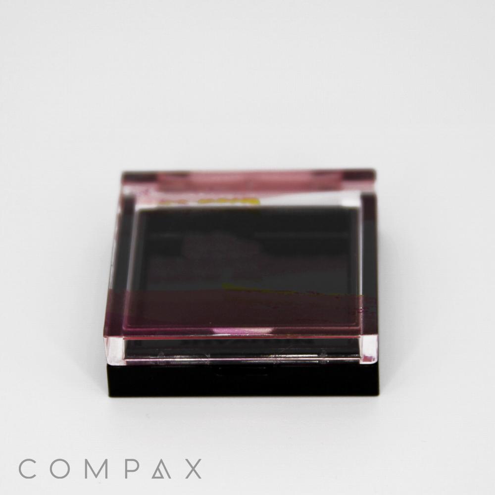 A closed Single Well Square Compact, featuring a black and transparent plastic design, reveals a partially visible pink substance inside. The brand name "COMPAX" is displayed at the bottom left corner of the image, set against a plain white background.