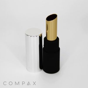 A cylindrical black Round Lipstick tube with a silver cap, positioned open. The lipstick inside is metallic gold. The background is plain white, and the brand name "COMPAX" is visible near the bottom left corner of the image.