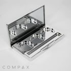 A silver-toned 3 Well Rectangular Compact with an open lid revealing a mirror and three circular compartments for holding makeup pans. A small applicator brush is positioned inside the case. The word "COMPAX" is visible on the surface below the compact.