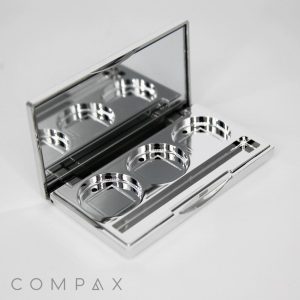 A silver-toned 3 Well Rectangular Compact with an open lid revealing a mirror and three circular compartments for holding makeup pans. A small applicator brush is positioned inside the case. The word "COMPAX" is visible on the surface below the compact.