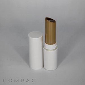 A gold cylindrical Slimline Round Lipstick with a white cap is positioned upright. The cap is removed and placed next to the container, revealing the golden lipstick inside. The background is plain and light-colored. The word "COMPAX" is visible in the lower left corner.