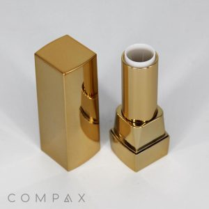 A Square Lipstick in a gold-colored tube, with its cap removed, is shown standing upright on a white surface. The brand name COMPAX is visible in the lower-left corner of the image. The lipstick itself appears unused and pristine, featuring a shiny, reflective finish.