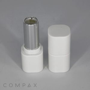A white square Stackable Twist-up Lipstick container with a matte finish sits open, revealing its metallic silver inner tube. The cap, also white and rectangular, is placed beside it. The background is plain and gray. The word "COMPAX" is visible at the bottom of the image.