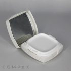 An open Square Cushion Compact in white displays an empty, square-shaped space for makeup alongside an integrated mirror on the inside of the lid. The compact is set against a neutral, light gray surface with "COMPAX" subtly visible at the bottom left corner of the image.