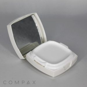 An open Square Cushion Compact in white displays an empty, square-shaped space for makeup alongside an integrated mirror on the inside of the lid. The compact is set against a neutral, light gray surface with "COMPAX" subtly visible at the bottom left corner of the image.