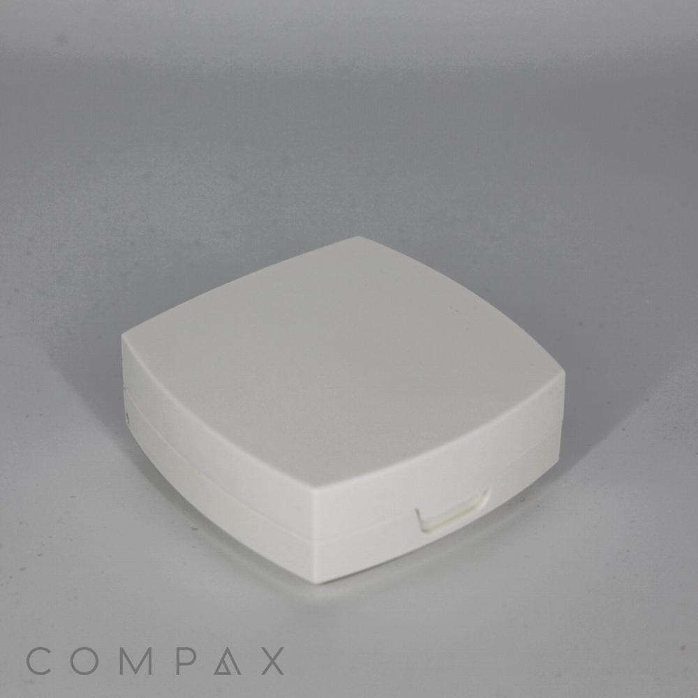 A small, white Square Cushion Compact with rounded edges and a small latch at the front. The background is a plain, light gray surface. The word "COMPAX" appears in the bottom left corner.
