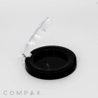 A black round compact case with a transparent lid standing open. The compact is empty, revealing the black interior. The brand name "COMPAX" is visible at the bottom left corner of the image.