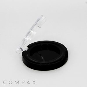 A black round compact case with a transparent lid standing open. The compact is empty, revealing the black interior. The brand name "COMPAX" is visible at the bottom left corner of the image.