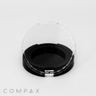 A round, black object with a transparent, semi-circular cover is displayed against a plain white background. The cover is open, revealing a black interior. The word "COMPAX" is printed in the bottom left corner.