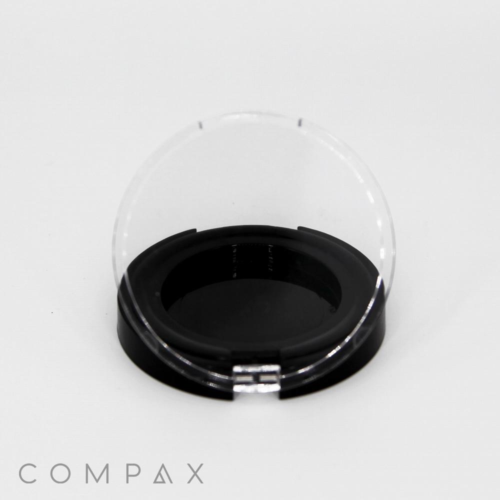 A round, black object with a transparent, semi-circular cover is displayed against a plain white background. The cover is open, revealing a black interior. The word "COMPAX" is printed in the bottom left corner.