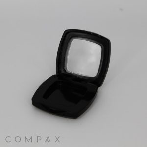 The image shows an empty, open black compact case with a small mirror inside. The case is square with rounded edges and has a glossy finish. The word "COMPAQ" is printed in light grey near the bottom left corner of the image.