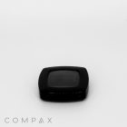 A black, square-shaped compact with a transparent lid rests on a white background. The word "COMPAX" is printed in light gray at the bottom left corner of the image.