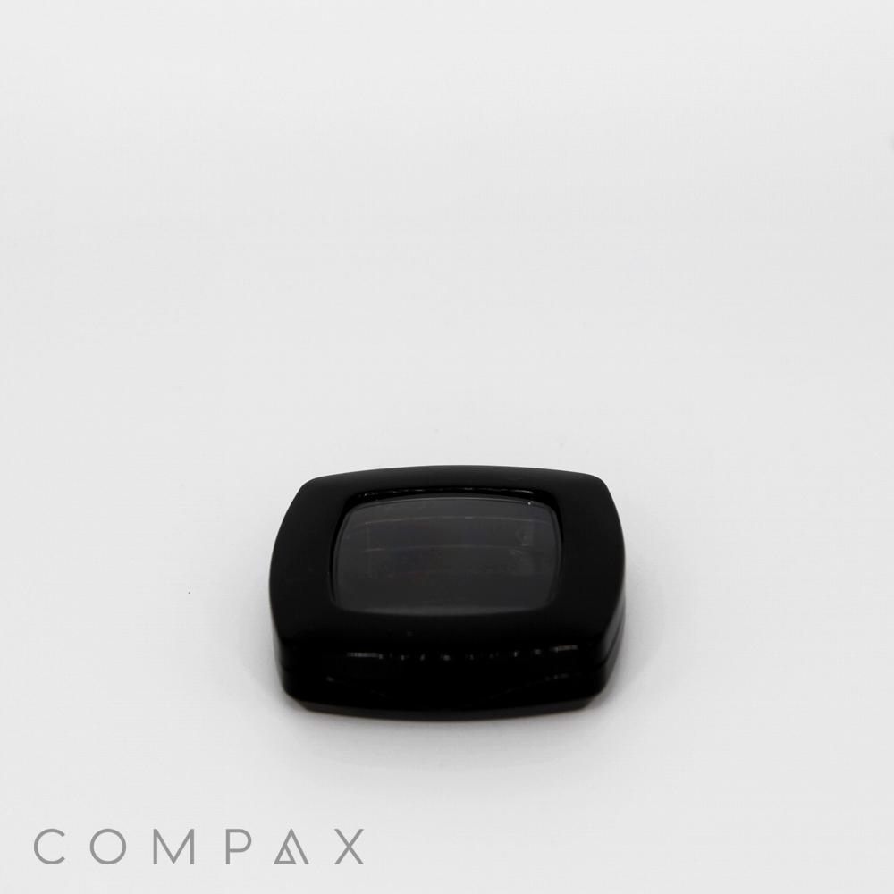 A black, square-shaped compact with a transparent lid rests on a white background. The word "COMPAX" is printed in light gray at the bottom left corner of the image.