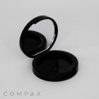 An open black compact case with a mirror on the upper lid and an empty compartment on the bottom. The background is plain white, and the word "COMPAX" is written in the lower left corner.