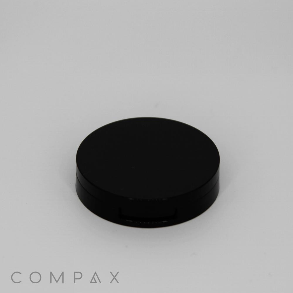 A black, round compact case with a smooth, matte finish is placed against a plain white background. The word "COMPAX" is printed in light gray, modern font in the bottom-left corner.