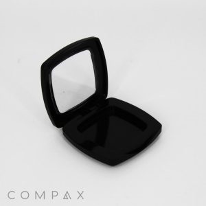 A black, square, empty compact case with a hinged lid partially open, revealing a mirror on the inside of the lid. The case is placed on a plain white background. The word "COMPAX" is written at the bottom left corner of the image.
