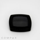 A square, black compact case with a slightly rounded edge is centered on a white background. The lens or cover appears to be slightly reflective. The word "COMPAX" is written in capital letters at the bottom left of the image.