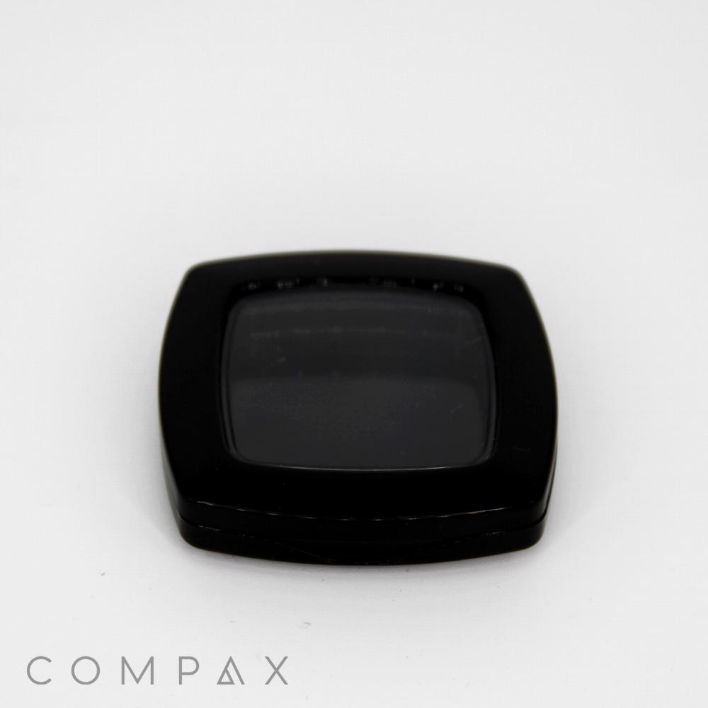 A square, black compact case with a slightly rounded edge is centered on a white background. The lens or cover appears to be slightly reflective. The word "COMPAX" is written in capital letters at the bottom left of the image.