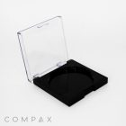 A square black makeup compact is open, revealing an empty circular compartment inside. The compact has a transparent lid, and the product branding "COMPAX" is visible at the bottom left corner of the image. The background is plain white.