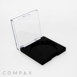 A square black makeup compact is open, revealing an empty circular compartment inside. The compact has a transparent lid, and the product branding "COMPAX" is visible at the bottom left corner of the image. The background is plain white.