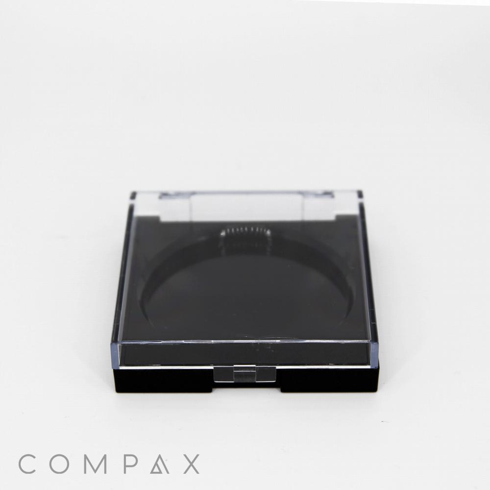A black compact case with a clear lid, bearing the brand name "COMPAX" at the bottom left corner. The inside of the case is empty, displaying a circular recess likely meant to hold a cosmetic product. The image background is plain white.