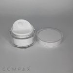 A small, clear plastic container of white cream with a white inner lid is displayed against a gray background. The main lid is placed next to the container. The brand name "COMPAX" is faintly visible in the bottom left corner.