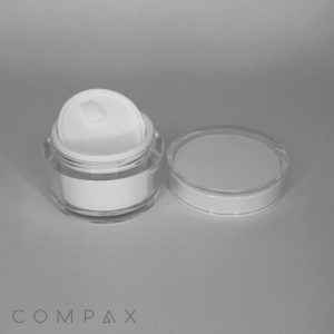 A small, clear plastic container of white cream with a white inner lid is displayed against a gray background. The main lid is placed next to the container. The brand name "COMPAX" is faintly visible in the bottom left corner.