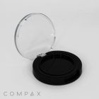 A round black compact case with a transparent lid is open. The interior is empty, designed to hold makeup such as powder or foundation. The brand name "COMPAX" is printed in gray letters in the bottom left corner of the image. The background is plain white.