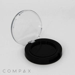 A round black compact case with a transparent lid is open. The interior is empty, designed to hold makeup such as powder or foundation. The brand name "COMPAX" is printed in gray letters in the bottom left corner of the image. The background is plain white.