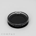 A black and clear round container sits on a light gray surface. The transparent lid reveals the inner black base. The word "COMPAX" is printed in light gray at the bottom left corner.