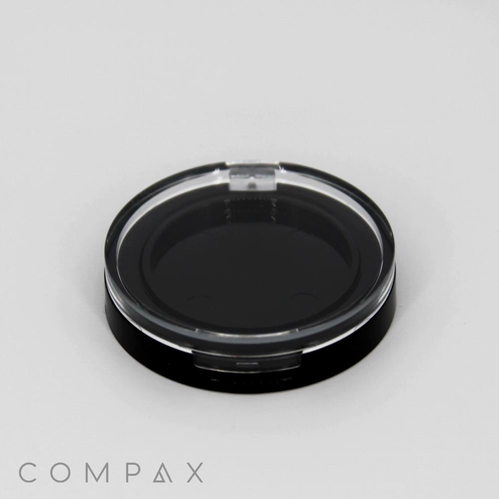 A black and clear round container sits on a light gray surface. The transparent lid reveals the inner black base. The word "COMPAX" is printed in light gray at the bottom left corner.