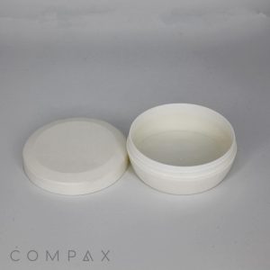 A small, round, white plastic container with a removable lid sits on a flat surface. The lid is placed to the left of the container, revealing its empty interior. The background is light gray, and the word "COMPAX" is visible at the bottom left corner.