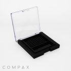 A small, square black compact with a clear lid, slightly open, displaying an empty grid-patterned compartment inside. The word "COMPAX" is written in light gray capital letters at the bottom left corner. The background is solid white.