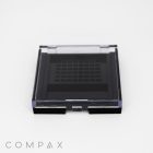 A black square compact case with a grid pattern on the inside of the lid, displayed on a white background. The word "COMPAX" is faintly visible at the bottom left corner.
