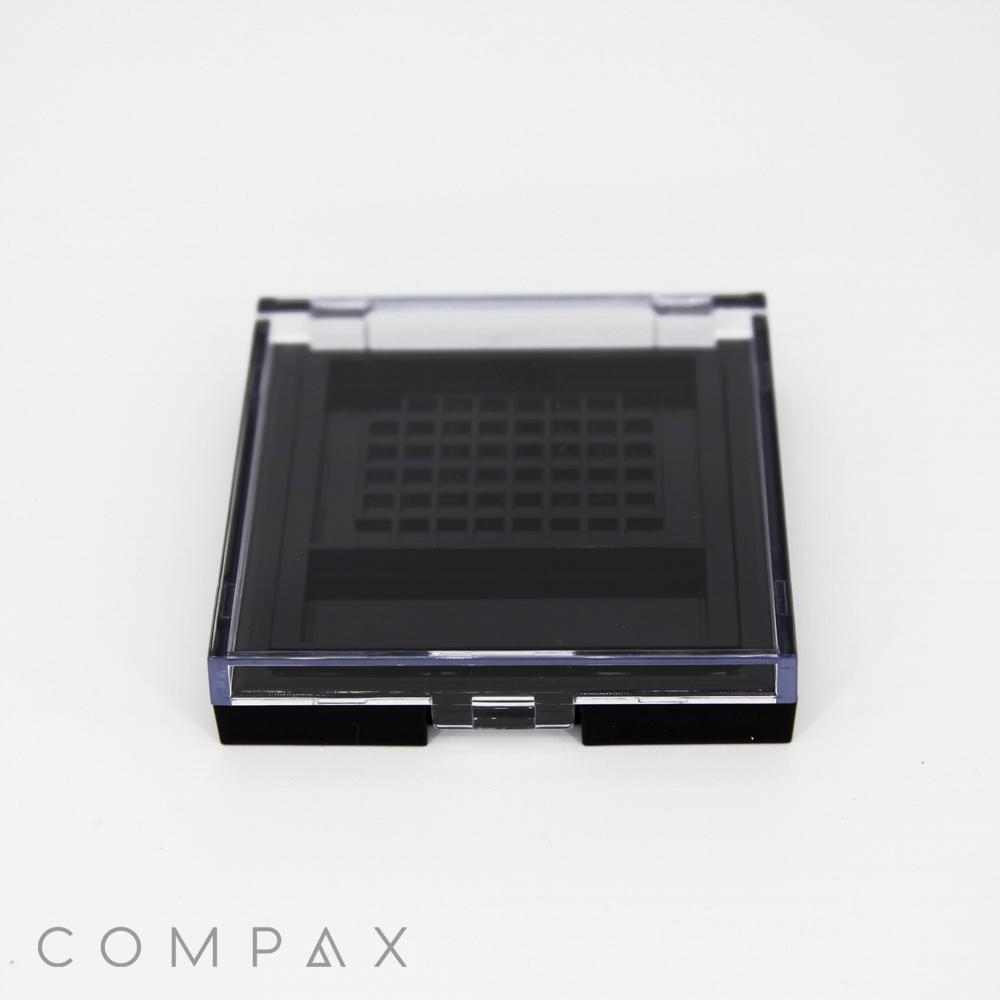 A black square compact case with a grid pattern on the inside of the lid, displayed on a white background. The word "COMPAX" is faintly visible at the bottom left corner.