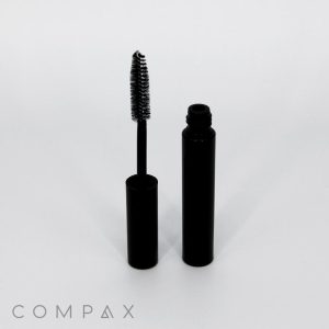A Round Mascara Tube in black, with the cap removed, stands upright on a white surface. The mascara wand, adorned with bristles coated in black mascara, leans gracefully against the tube. In the lower left corner, the word "COMPAX" is printed in gray letters.