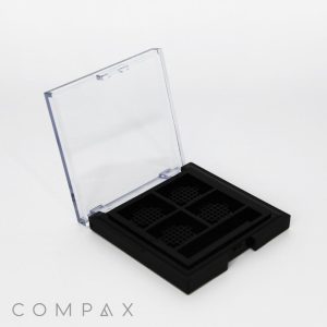 A black compact case with a transparent lid is displayed. The interior features four square compartments, each with a grid-like textured pattern. The word "COMPAX" is printed on the bottom left of the image.