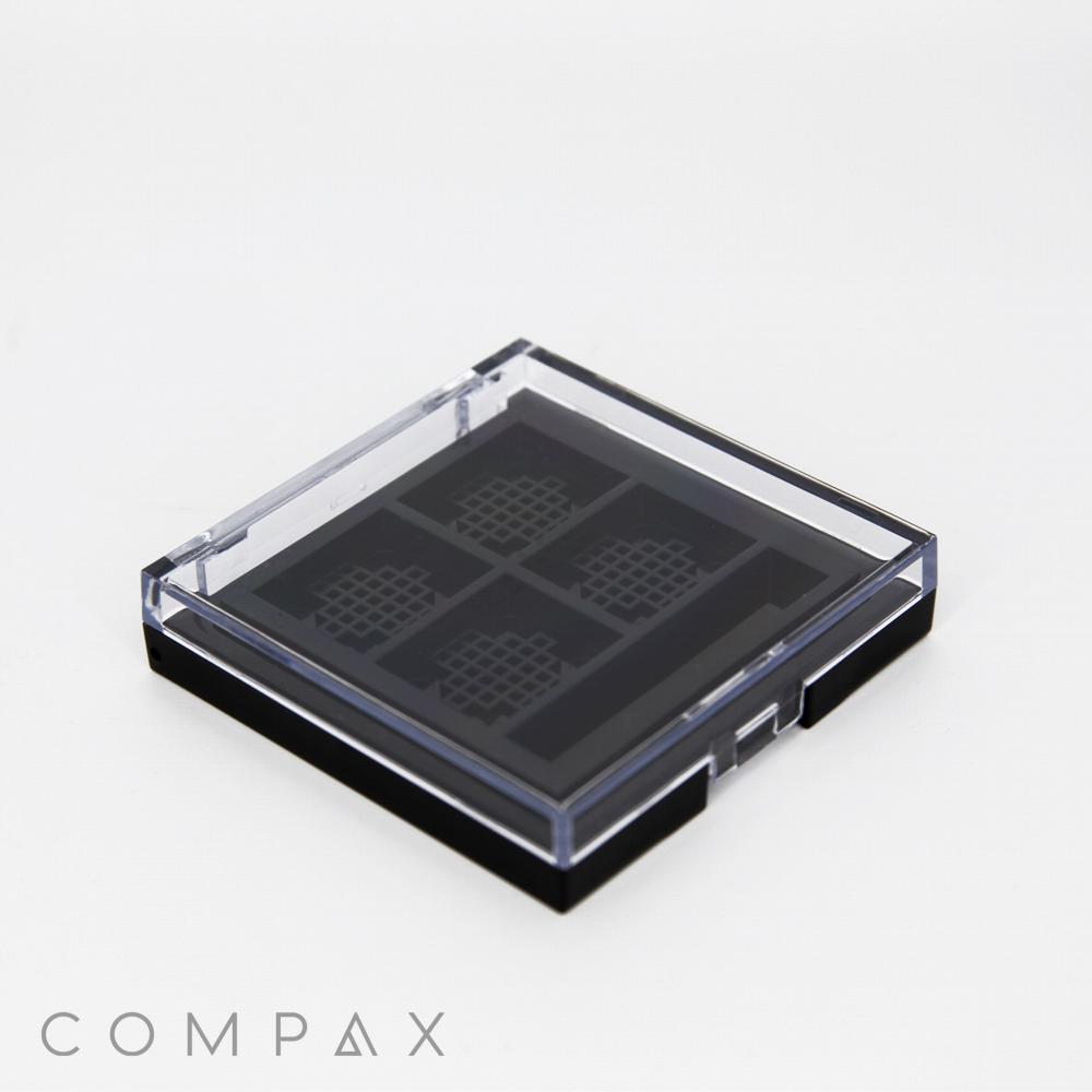 A square compact makeup case with a clear lid, displaying four evenly spaced compartments inside. The lid has a grid pattern, and the case is closed. The word "COMPAX" is printed in gray at the bottom left corner. The background is white.