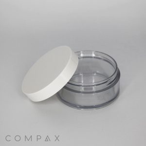 A clear, empty plastic container with a white short cap placed slightly ajar. The container is cylindrical and transparent, allowing the interior to be visible. The brand name "COMPAX" appears in lowercase letters at the bottom left corner of the image.