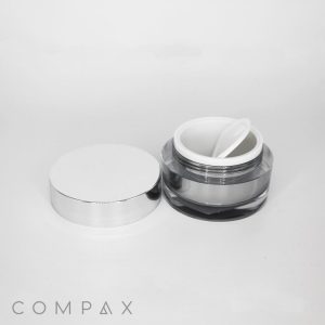 A small, round double-walled cosmetic jar with a metallic silver lid. The lid is placed beside the open jar, revealing a white inner container and a small plastic spatula resting on top. The jar is placed against a plain white background, and the brand name "COMPAX" is visible at the bottom left.