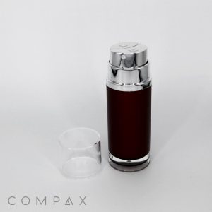 A metallic cylindrical container named "Round Dual Chamber Airless Bottle" with a shiny silver top and a transparent plastic cap placed beside it, featuring a dark brown body labeled "COMPAX" at the bottom left corner of the image. The background is plain white.