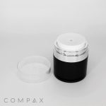 A cylindrical Round Airless Pump Jar in black and white with a transparent lid rests on a white surface. The lid is placed beside the jar. The brand name "COMPAX" is visible in the bottom left corner of the image.