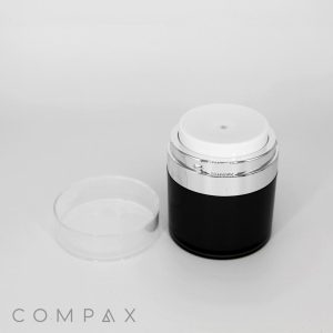 A cylindrical Round Airless Pump Jar in black and white with a transparent lid rests on a white surface. The lid is placed beside the jar. The brand name "COMPAX" is visible in the bottom left corner of the image.